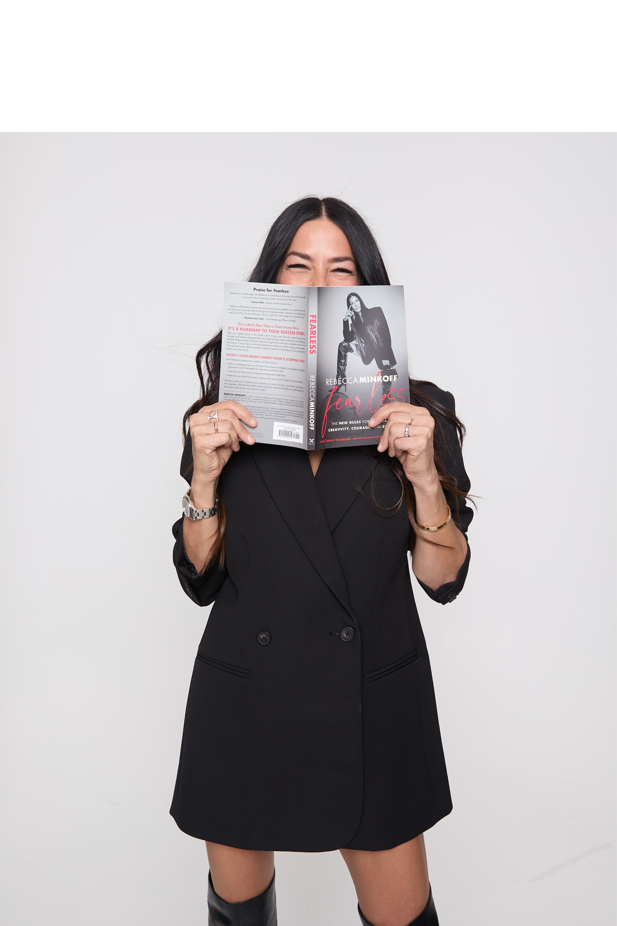 Woman wearing black dress, high boots holding a book in front of her face.