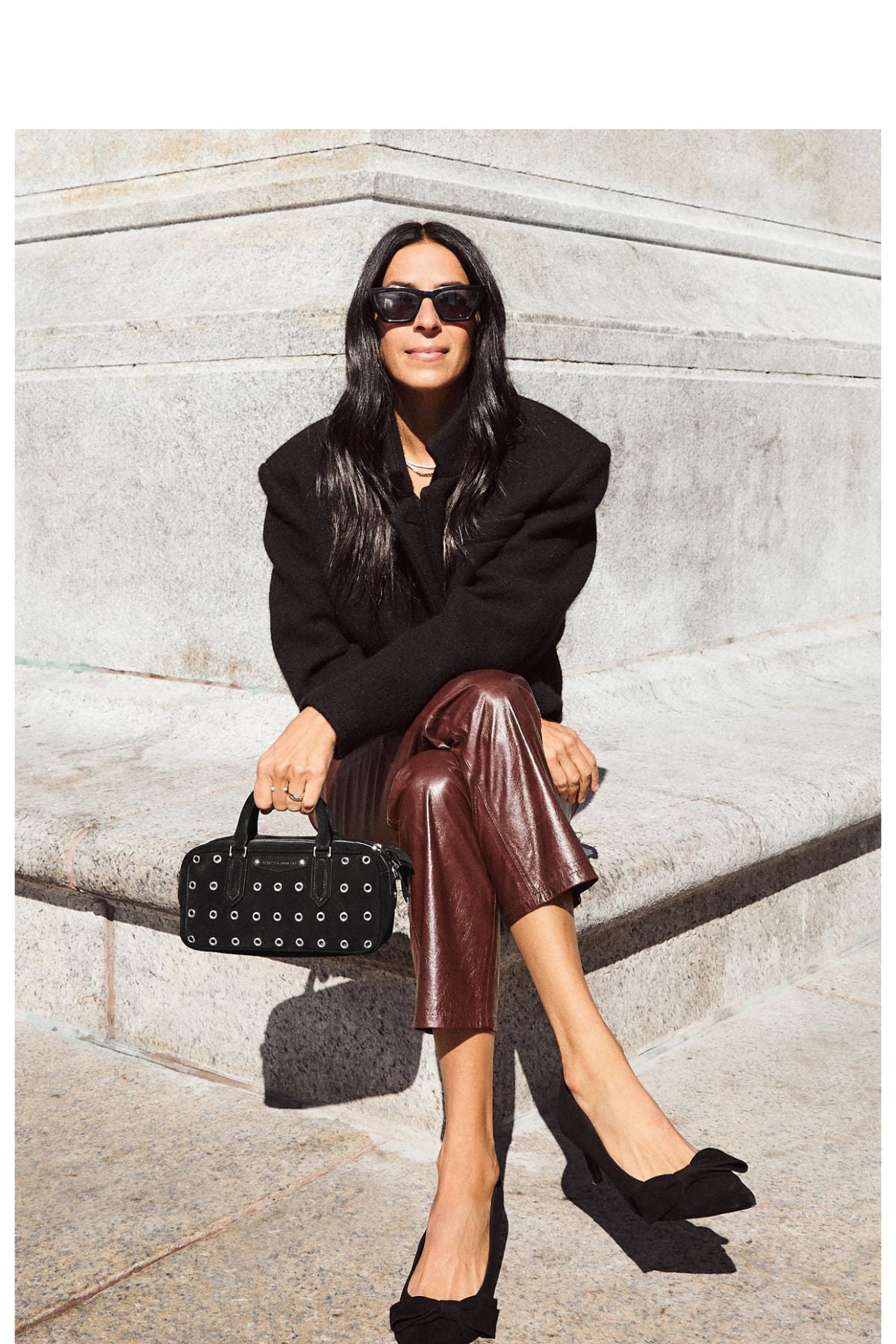 Woman in black sweater and brown leather pants sitting on a stone wall.